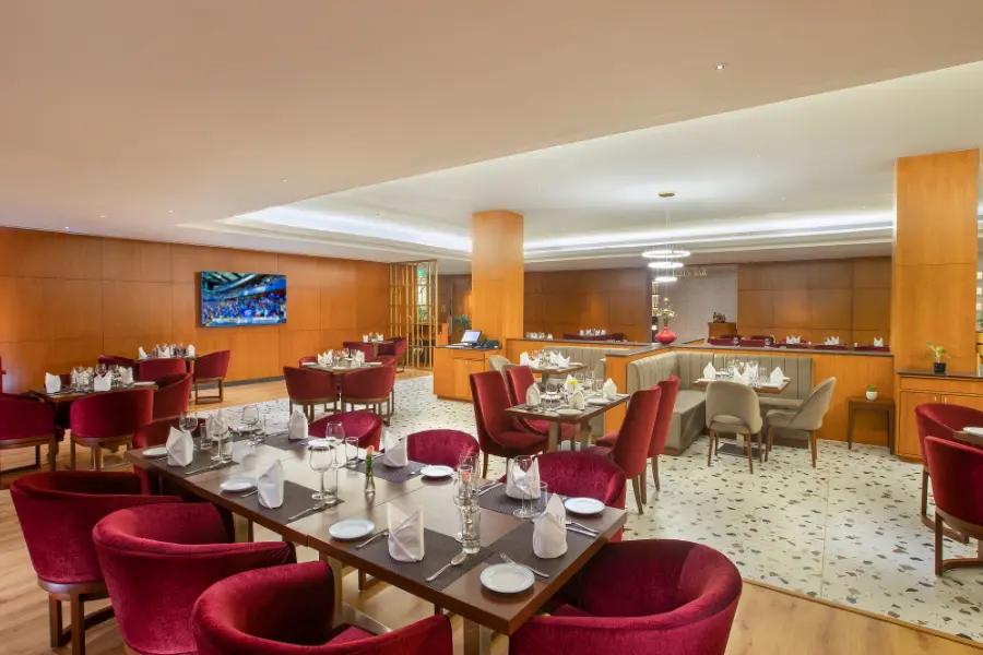 A hotel restaurant dining space with a clean, organized table setup. Tables are arranged with plates, glassware, and folded napkins. Seating includes red cushioned chairs. The flooring is patterned with small tile-style designs and the walls have wooden panels. The overall lighting is soft and warm, giving a premium dining feel.