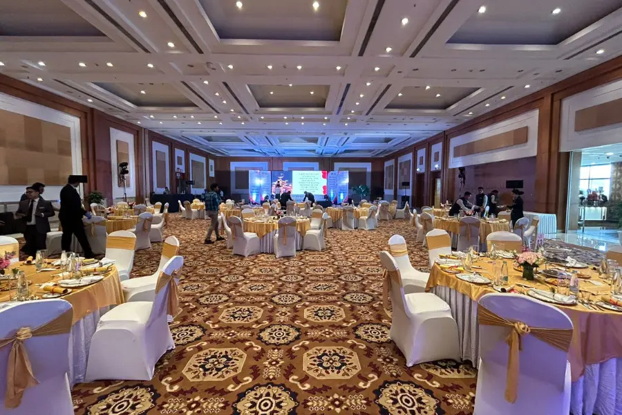 Elegant banquet hall set with round dining tables, white chairs with gold ribbons, and stage lighting for an event.