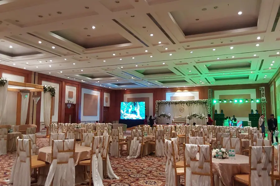Spacious indoor banquet hall with rows of chairs covered in light fabric, round tables, a decorated wedding stage with draped backdrop, and green accent lighting