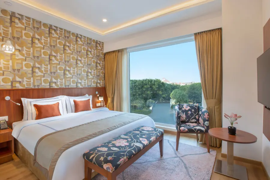 Hotel bedroom suite with a large bed, patterned accent wall, cushioned seating bench, one side chair, a small round table, wall-mounted TV, curtains, and a wide window view