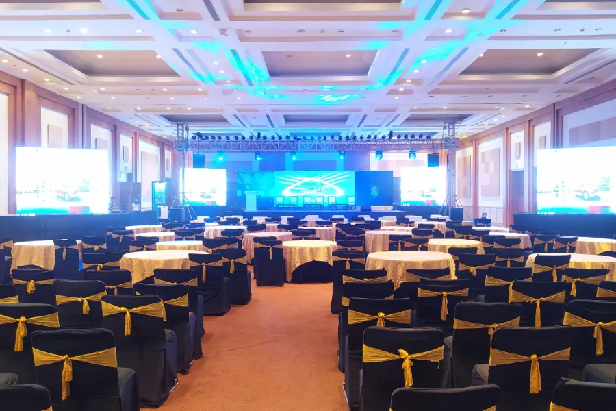 Large indoor convention hall with a stage, LED screens, ceiling lights, and multiple round tables with chairs arranged for an event
